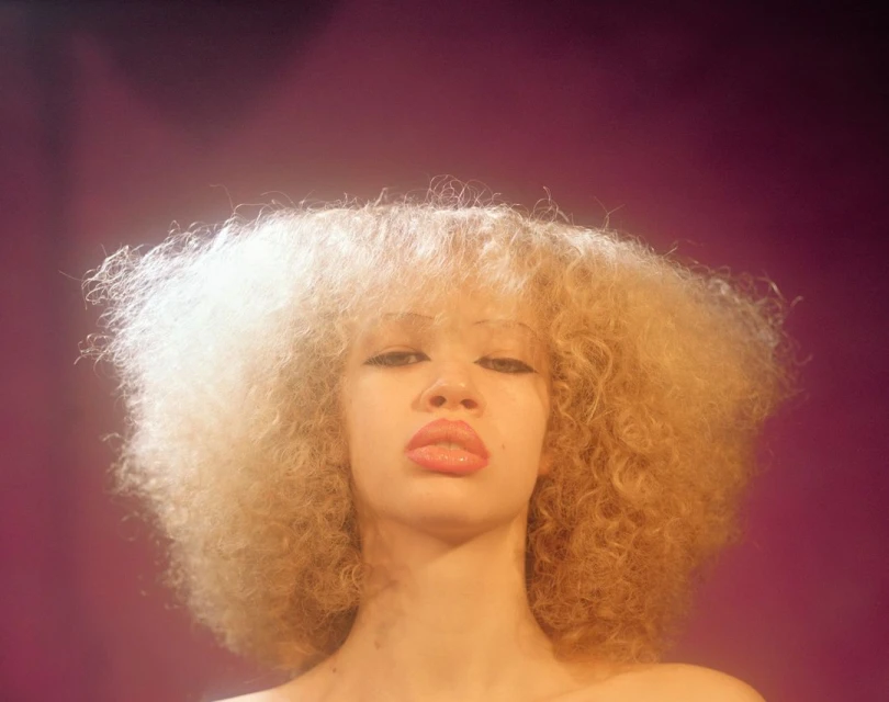 Portrait of a young adult female with light blonde curly hair and neutral expression in front of a soft pink background.