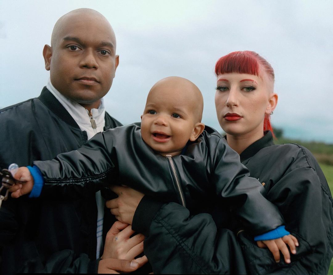 Portrait of a family of three outside in a field with a dark-haired man, fair-skinned woman with red hair, and a young child.