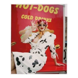 Woman in cow-print dress and headscarf posing before a red hot dog stand sign