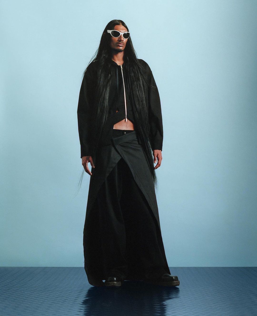 Fashion model wearing a black jacket, wrap skirt, and white sunglasses stands in front of a blue backdrop in a studio setting.