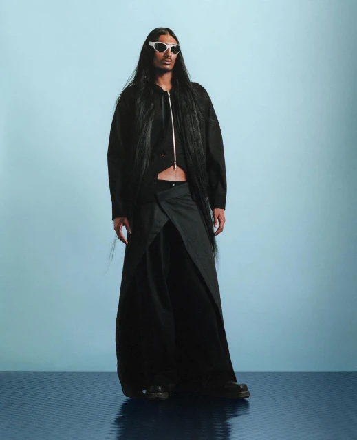 Fashion model wearing a black jacket, wrap skirt, and white sunglasses stands in front of a blue backdrop in a studio setting.