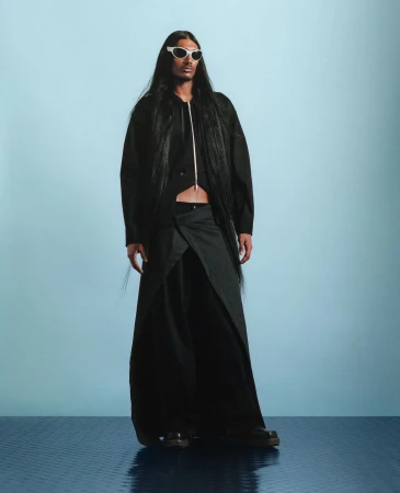 Fashion model wearing a black jacket, wrap skirt, and white sunglasses stands in front of a blue backdrop in a studio setting.