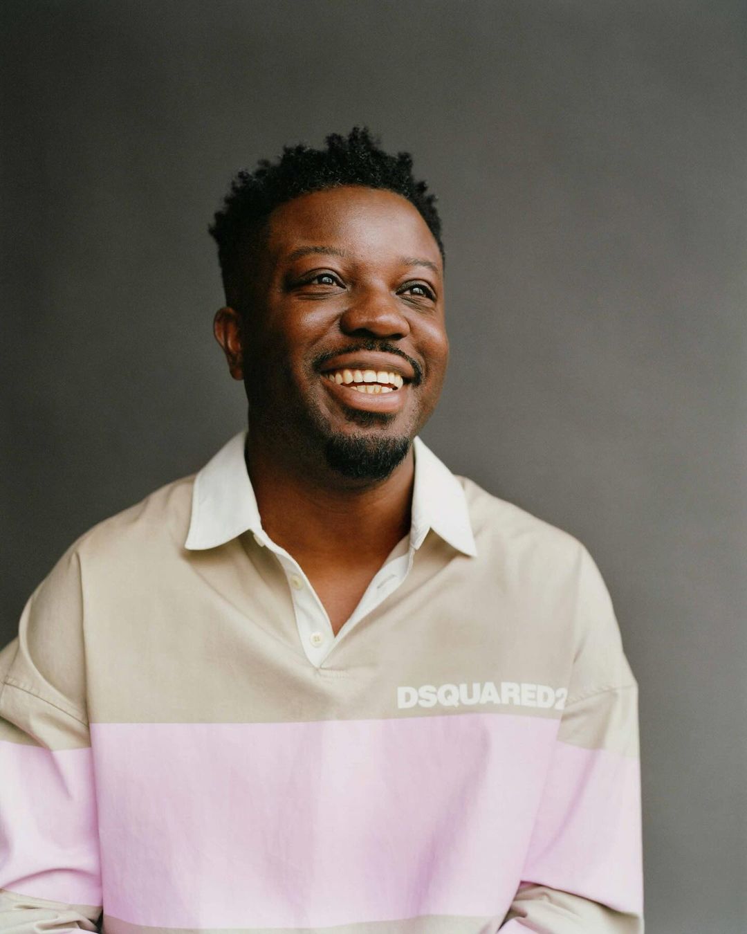 Portrait of a smiling African American man wearing a pink and beige polo shirt against a gray background. He is looking off camera.