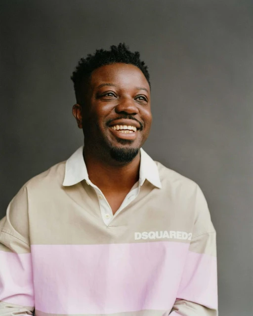 Portrait of a smiling African American man wearing a pink and beige polo shirt against a gray background. He is looking off camera.
