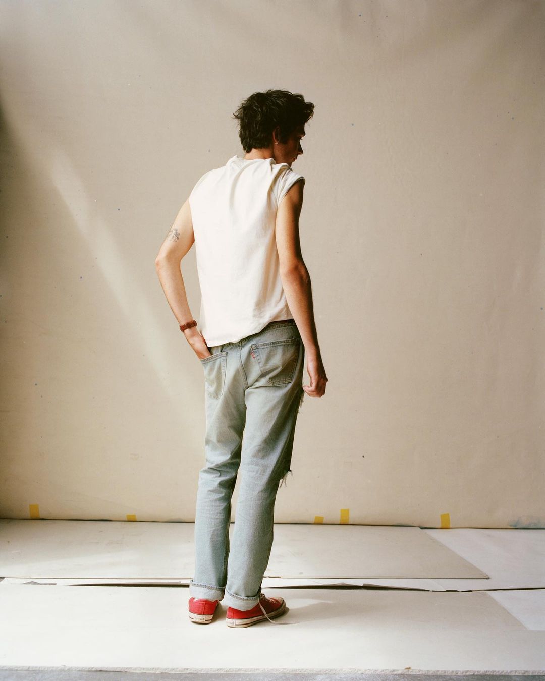 A stylish young adult stands in a studio with ripped jeans and a white sleeveless shirt, illuminated by natural light, creating a casual yet chic look.