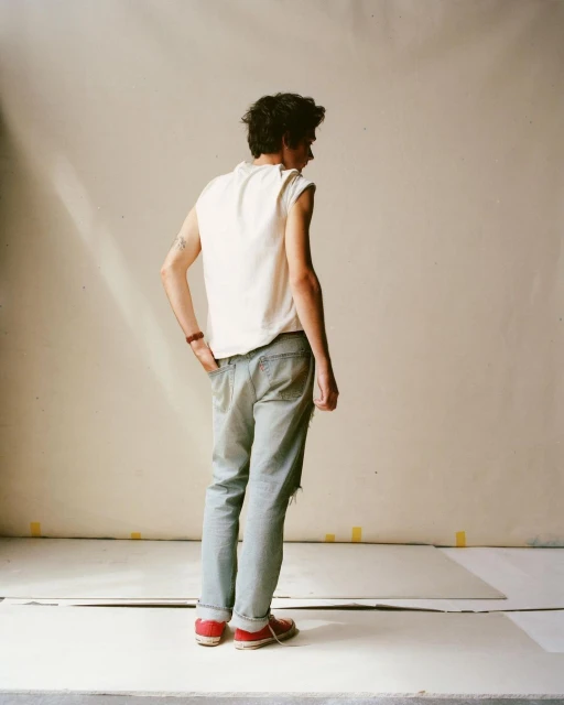 A stylish young adult stands in a studio with ripped jeans and a white sleeveless shirt, illuminated by natural light, creating a casual yet chic look.