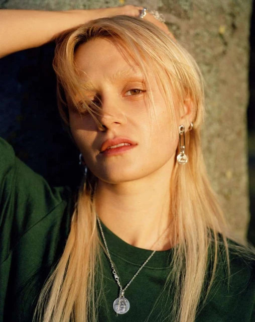 Portrait of a young blonde woman in a green shirt, showcasing silver earrings and a necklace against a blurred outdoor backdrop.