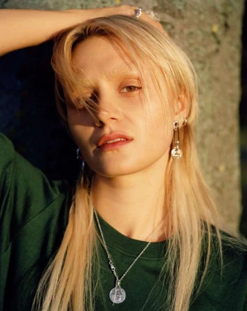 Portrait of a young blonde woman in a green shirt, showcasing silver earrings and a necklace against a blurred outdoor backdrop.