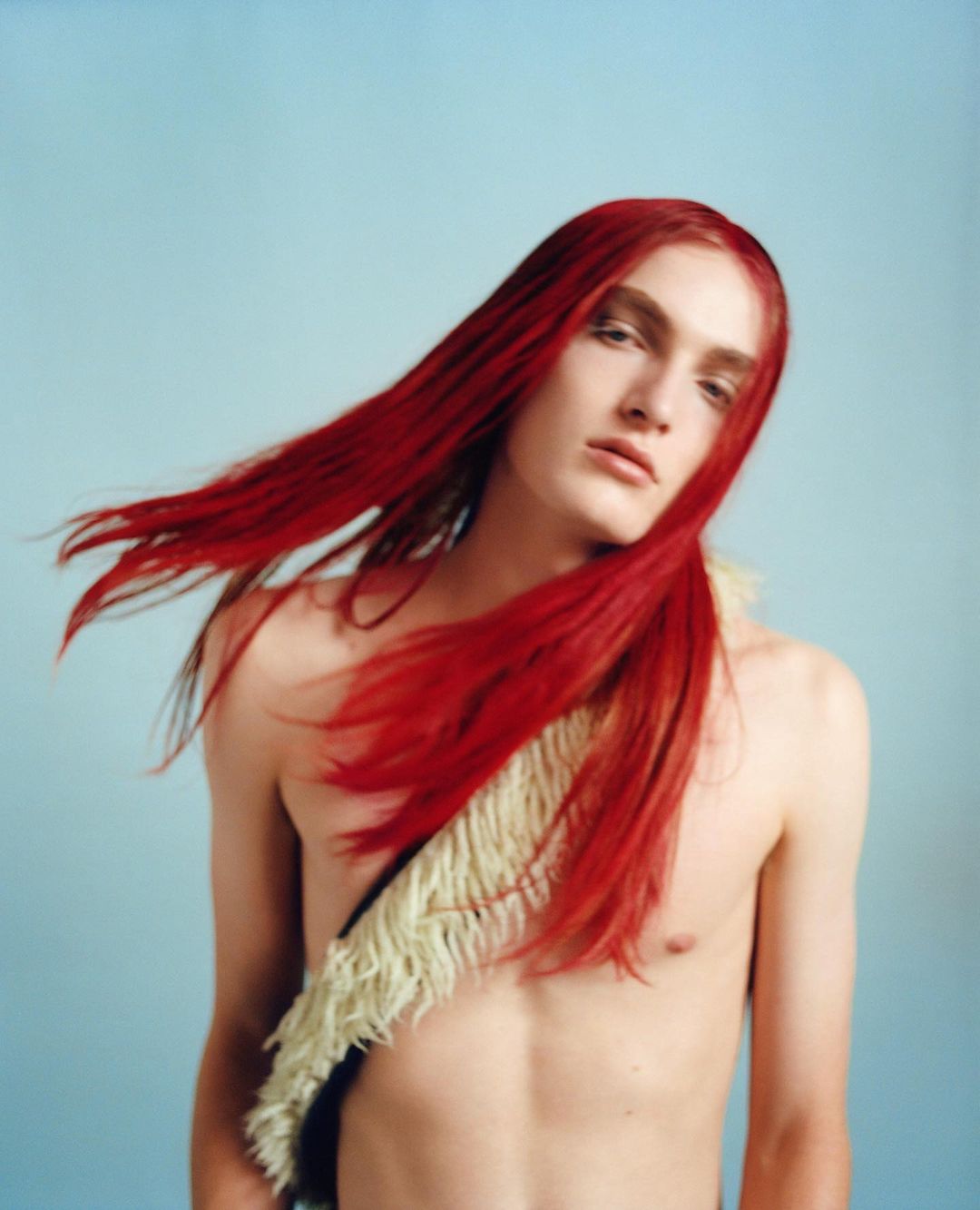 Fashion portrait of a shirtless adult with red hair, wearing a fur shawl against a pale blue background. Studio lighting.