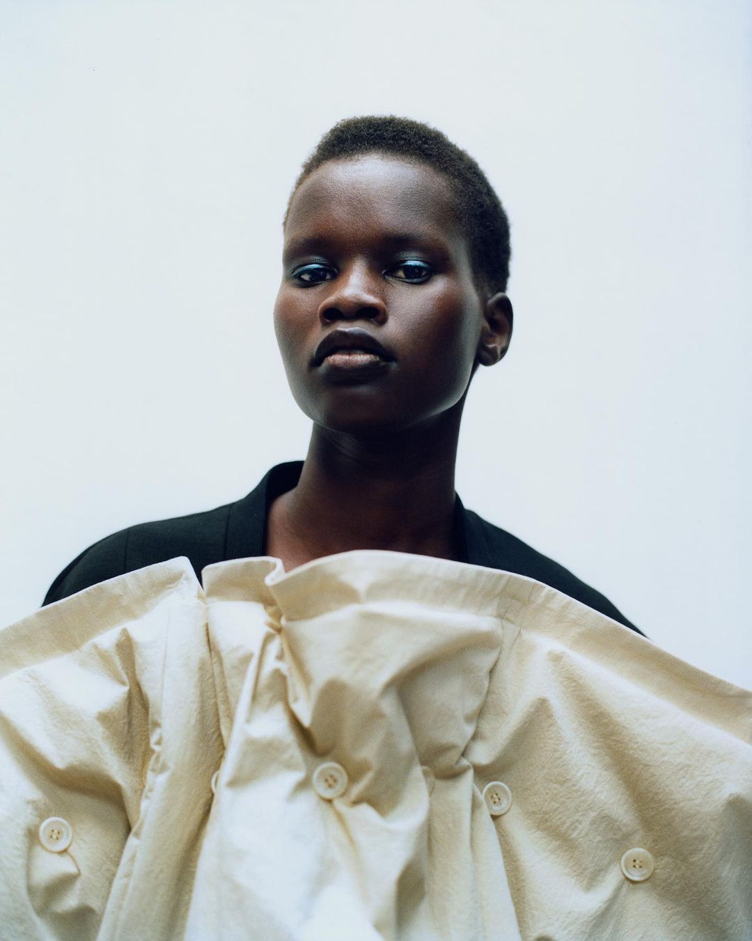 Close-up studio portrait of an African-descent adult female model in neutral makeup, wearing a black jacket and a large sculptural cream-colored top.