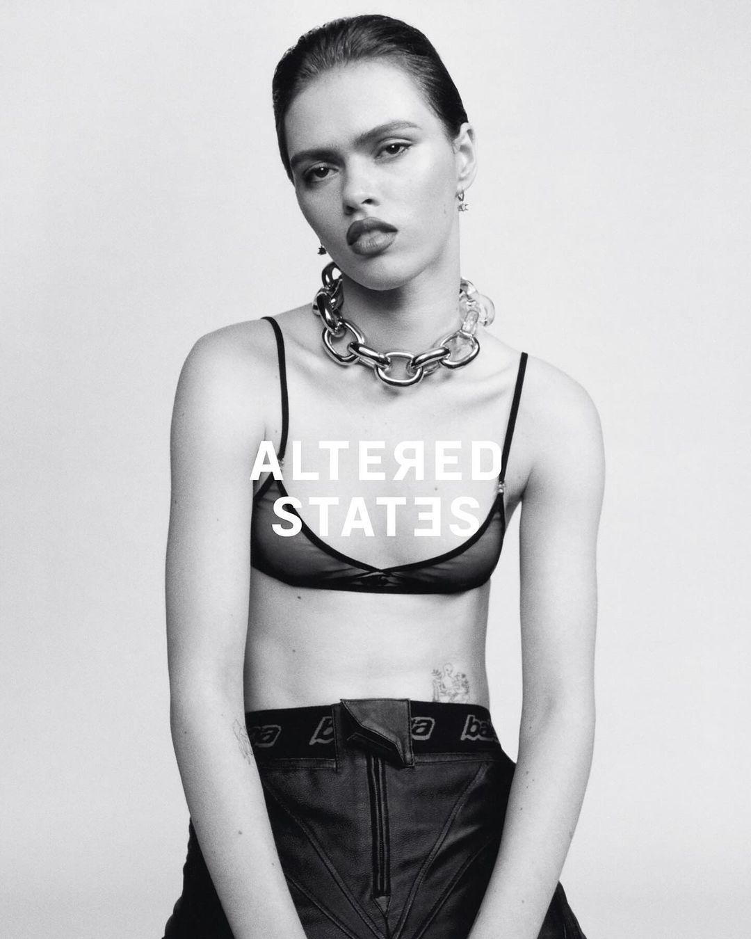 Black and white portrait of a woman wearing a bra top, pants and a chain necklace in a photo studio with white background.
