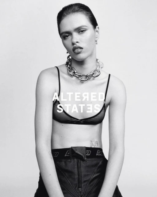 Black and white portrait of a woman wearing a bra top, pants and a chain necklace in a photo studio with white background.