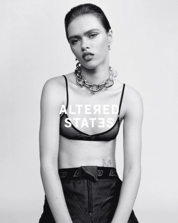 Black and white portrait of a woman wearing a bra top, pants and a chain necklace in a photo studio with white background.