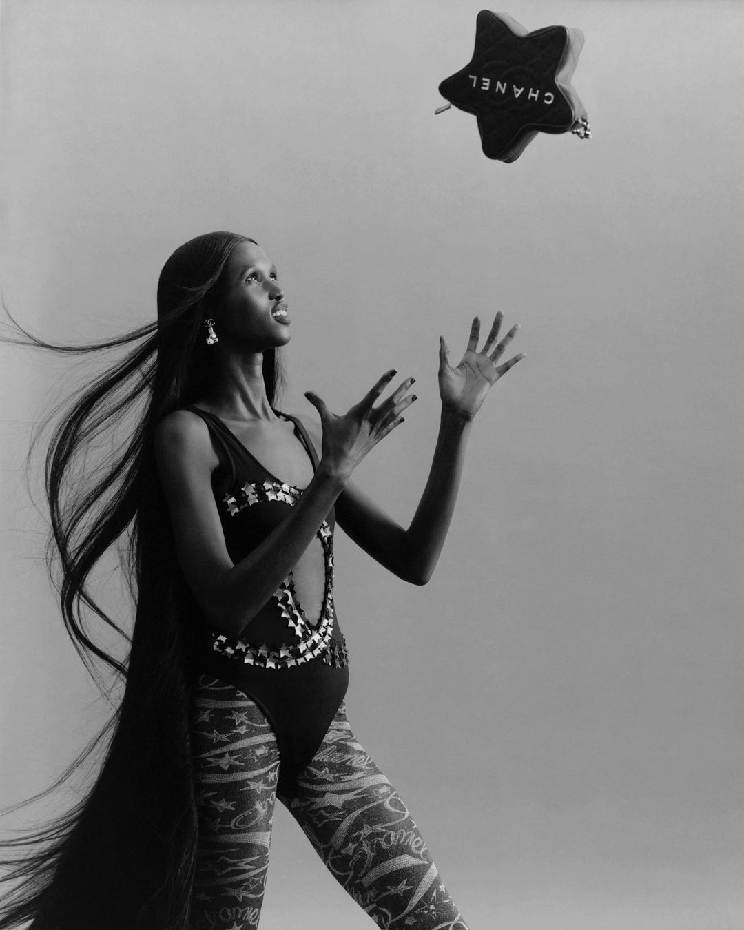 Black and white studio portrait of a model in patterned bodysuit catching a star-shaped Chanel bag against a graduated grey backdrop.