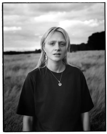 Portrait of a blonde woman wearing a dark t-shirt and pendant necklace outdoors in a field under a cloudy sky.