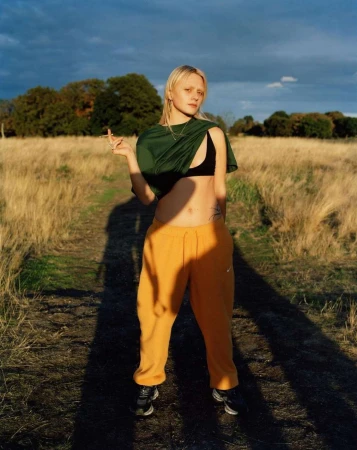 Blonde woman wearing a green t-shirt, black bralette, and yellow sweatpants standing in a grassy field in London.