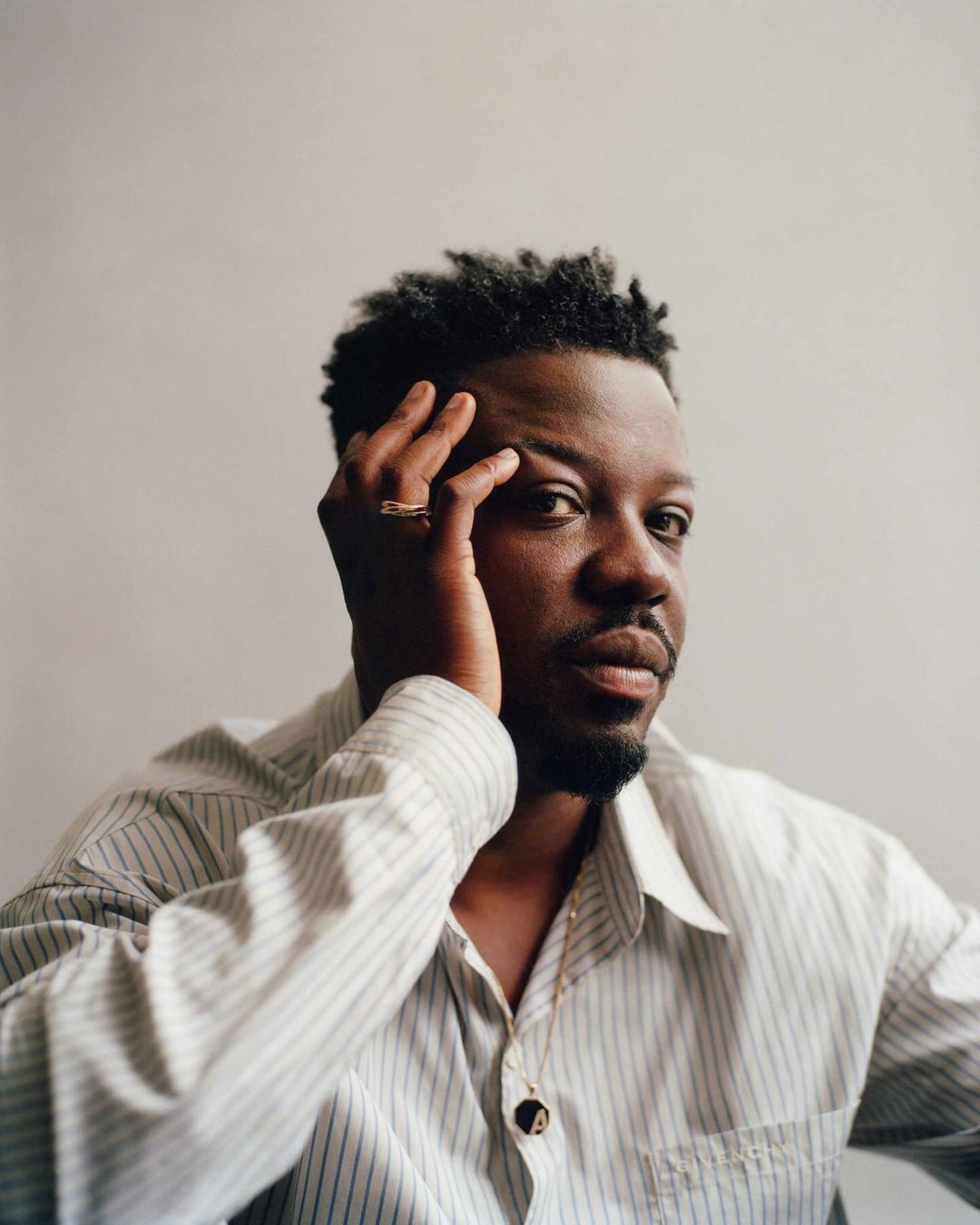 Portrait of an adult male with hand near his face, wearing a striped shirt and necklace, set against a neutral grey studio backdrop.
