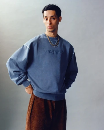 Studio shot of a young adult male model wearing a blue sweater and brown corduroy pants, posing with hands on hips and layered gold necklaces.