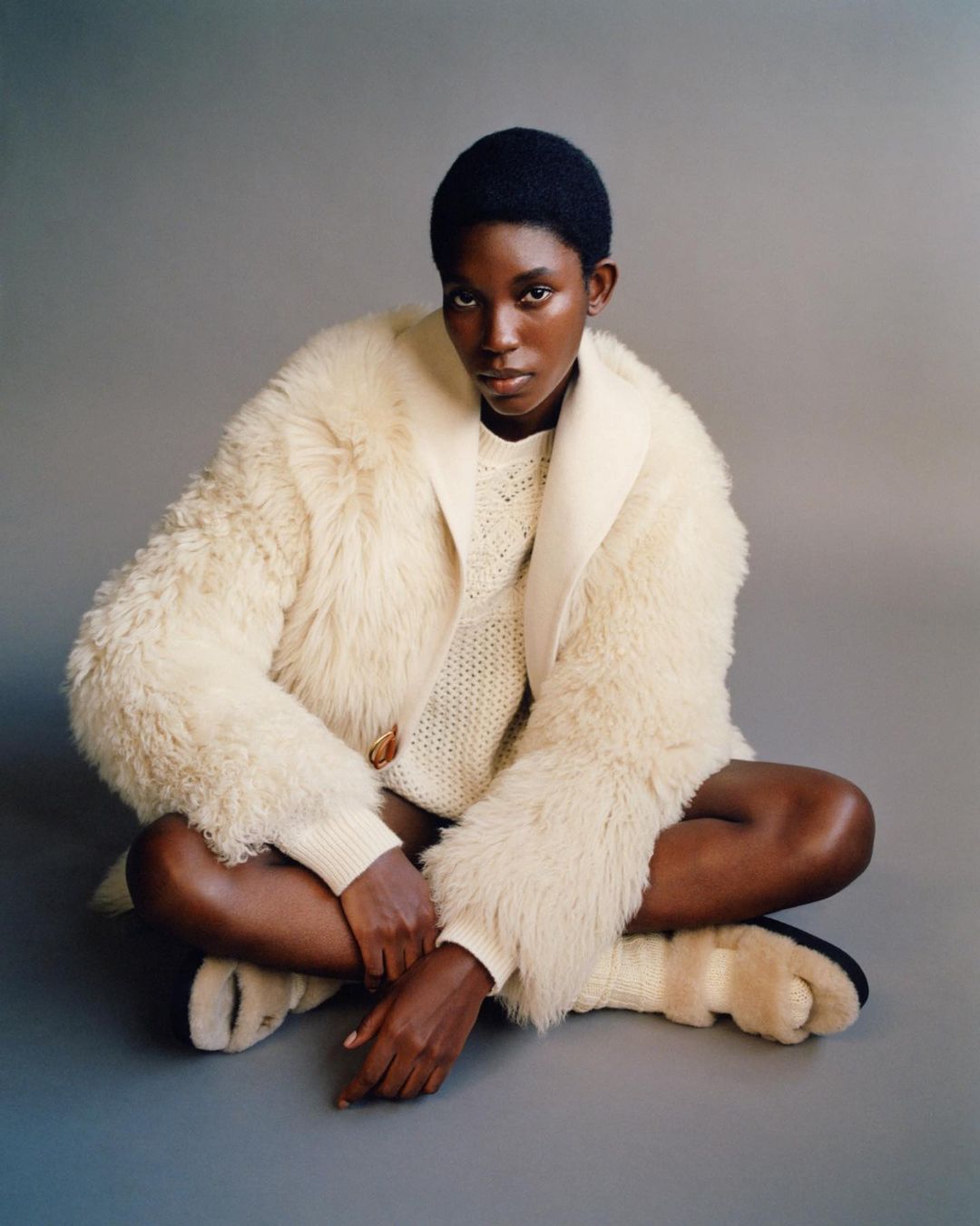 Adult female in a fur coat and sweater sits cross-legged on a grey studio backdrop, gazing directly at the camera in a fashion shot.