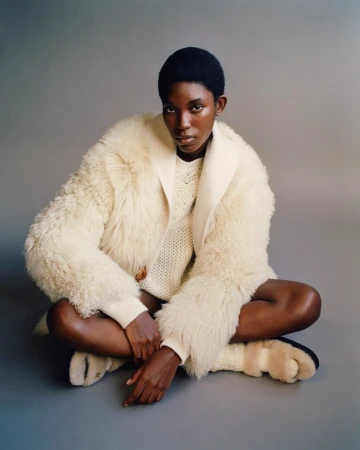 Adult female in a fur coat and sweater sits cross-legged on a grey studio backdrop, gazing directly at the camera in a fashion shot.