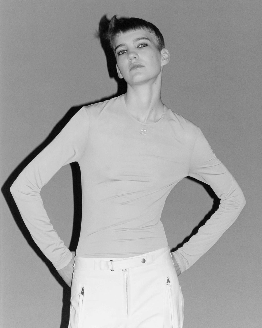 Monochrome portrait of a short-haired woman in a long-sleeve top and trousers posing against a grey backdrop by Lola and Pani.