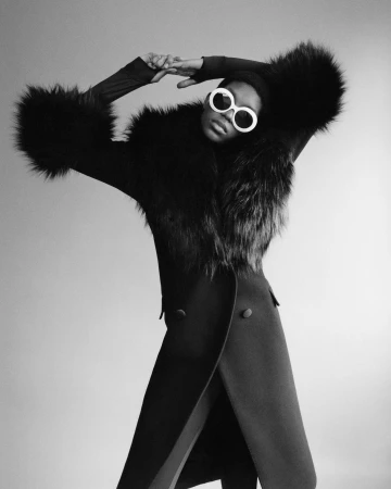 Model in white sunglasses and black fur-trimmed coat posing with arms raised against a plain light studio background.