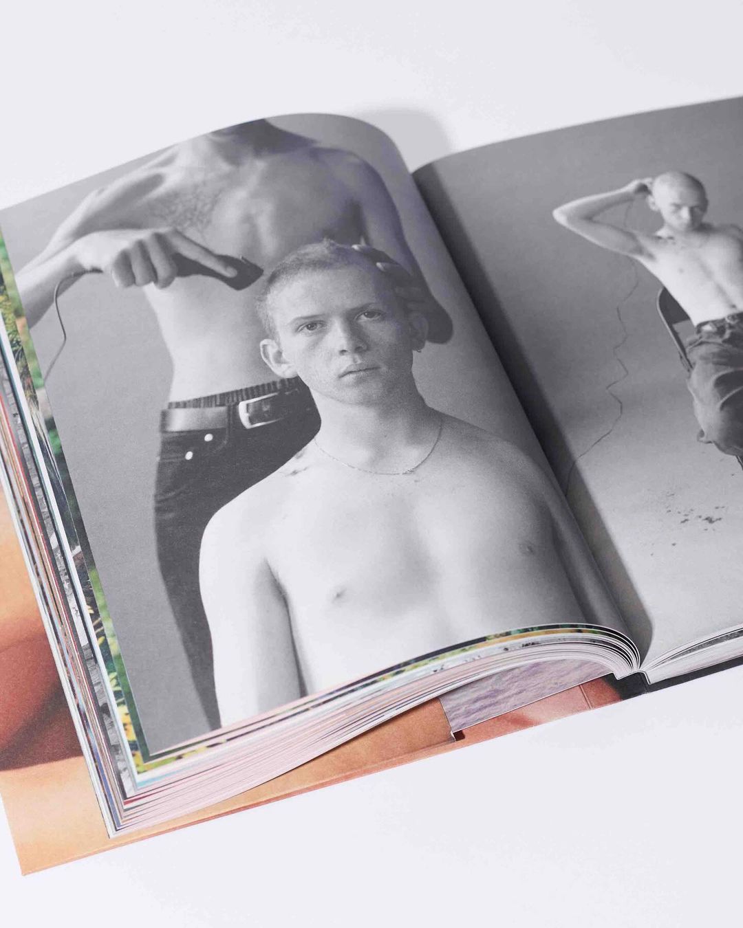 Open magazine features a black-and-white photo of shirtless men, one receiving a haircut, on a white surface with colorful page edges.