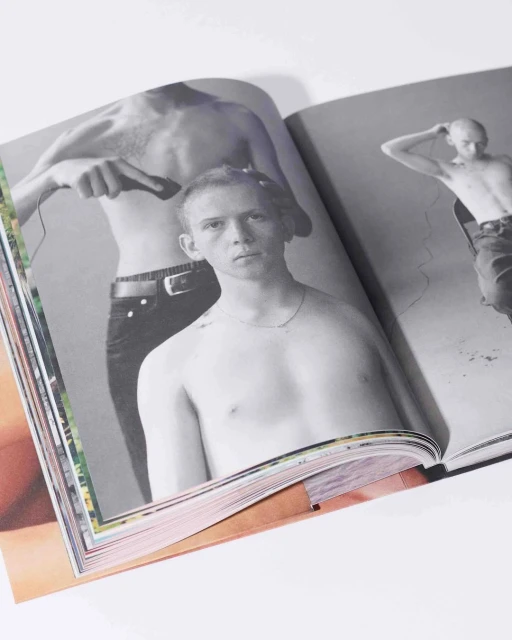 Open magazine features a black-and-white photo of shirtless men, one receiving a haircut, on a white surface with colorful page edges.
