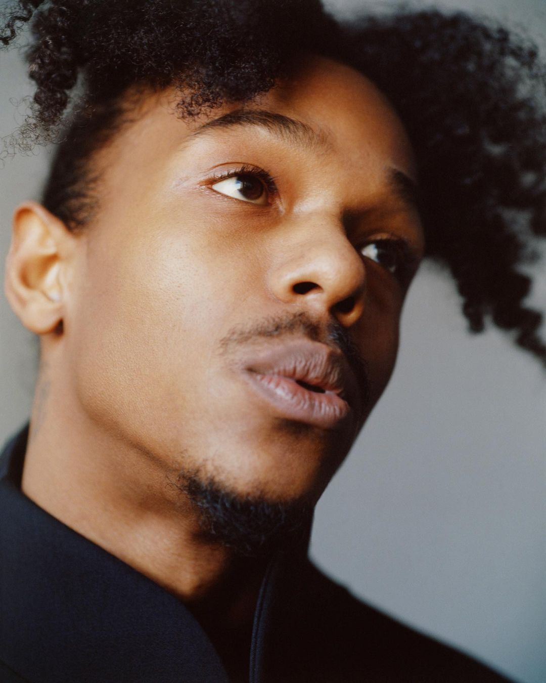 Close-up portrait of a handsome adult Black male with short curly hair, wearing a black jacket, gazing upward in an indoor setting.