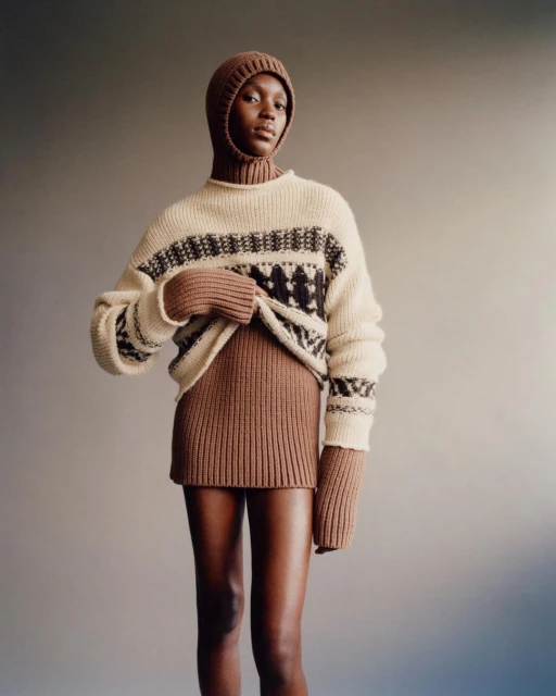 Fashionable woman in a patterned knit sweater and matching balaclava, posing in a studio with neutral background. Warm-toned color grading.