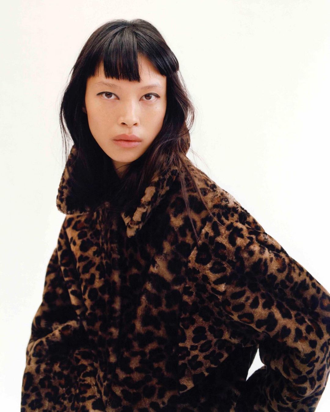 Fashion portrait of an Asian woman with dark hair wearing a leopard print coat against a plain white backdrop in a photo studio.
