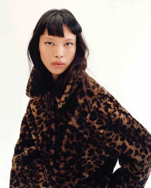 Fashion portrait of an Asian woman with dark hair wearing a leopard print coat against a plain white backdrop in a photo studio.
