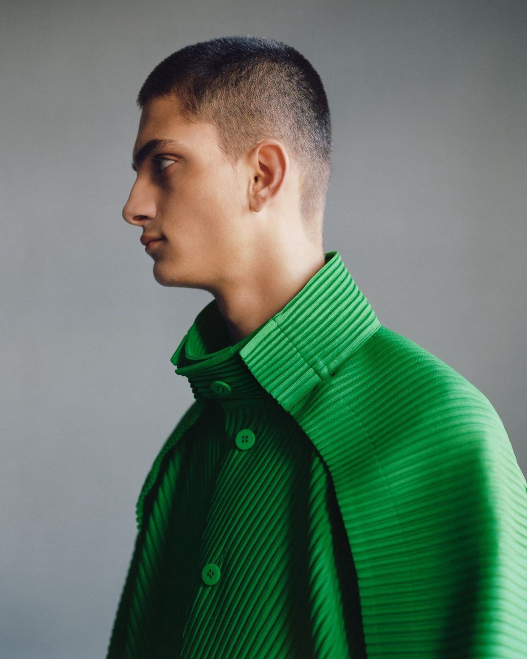 Profile portrait of a young adult male with short hair wearing a green pleated shirt against a grey background in a studio setting.