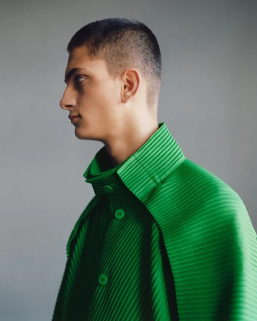 Profile portrait of a young adult male with short hair wearing a green pleated shirt against a grey background in a studio setting.