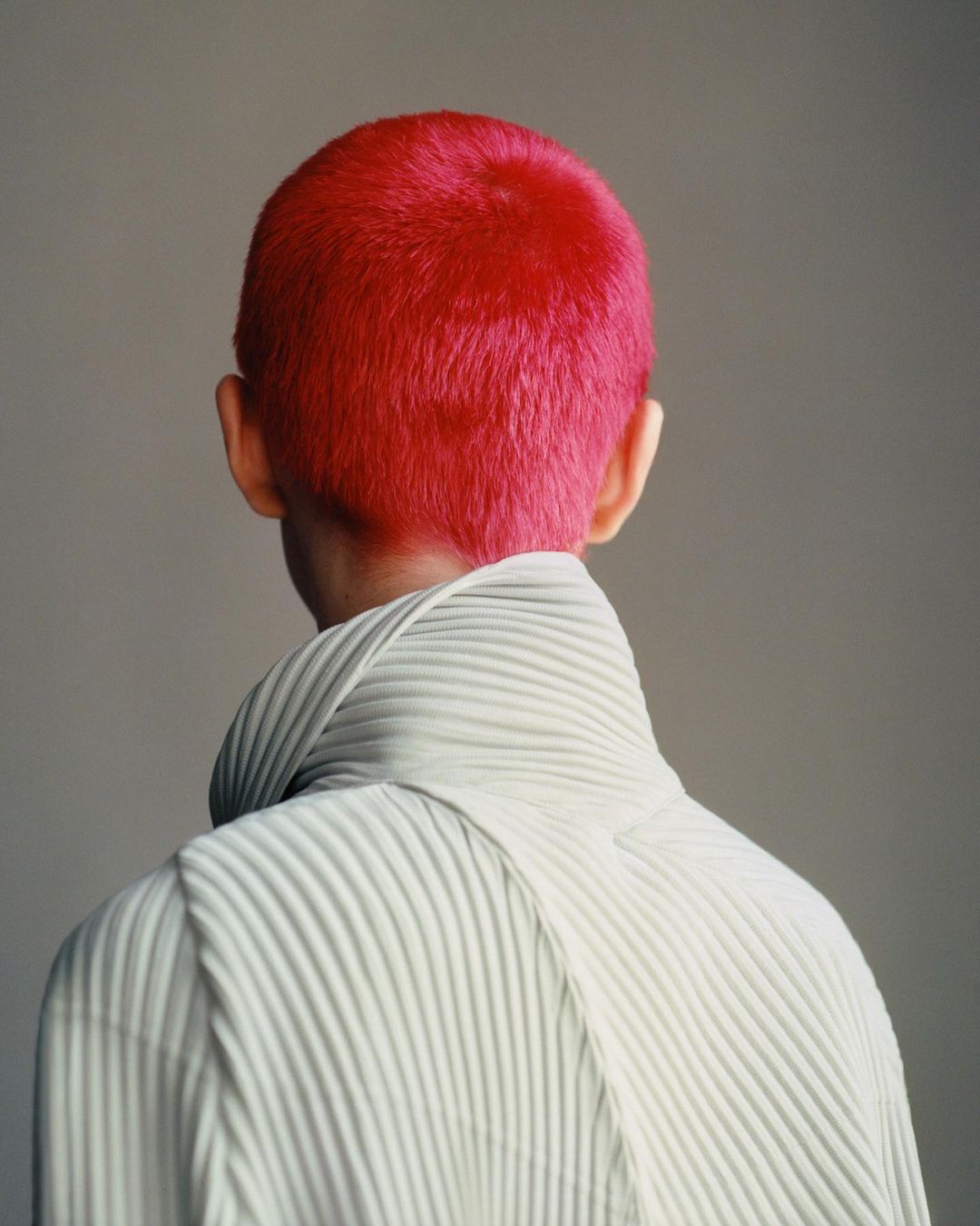 Close-up of person with a vibrant pink buzzcut, wearing a white high-necked, vertically-pleated top against a neutral backdrop.