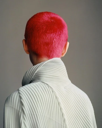Close-up of person with a vibrant pink buzzcut, wearing a white high-necked, vertically-pleated top against a neutral backdrop.