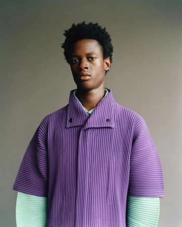 Portrait of a young Black man wearing a vibrant purple, collared shirt, posing against a grey backdrop in a studio setting.