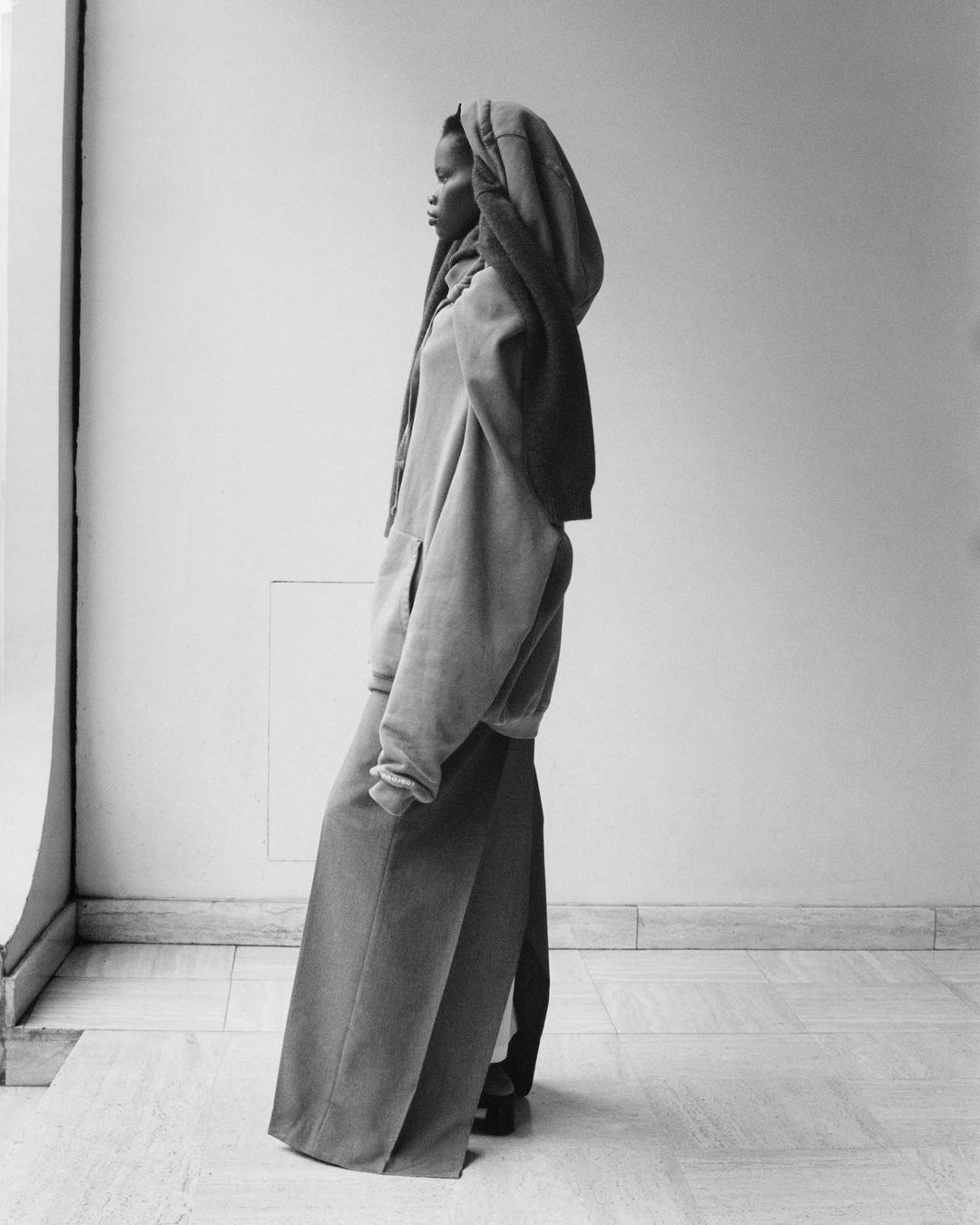 Fashion shot of a Black female model in a full-length, oversized grey hoodie dress, posing in profile against a white wall with window light.
