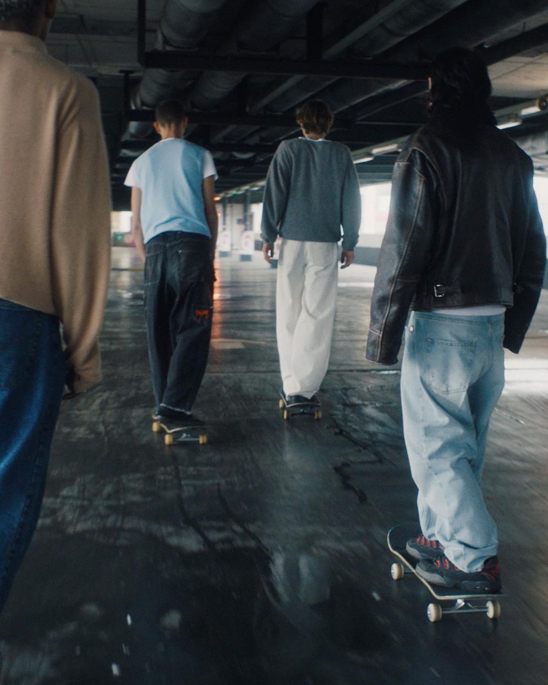 Group of teens skateboard on a wet, urban surface in an underpass, showing casual clothing, skateboards, and movement with a wide perspective.