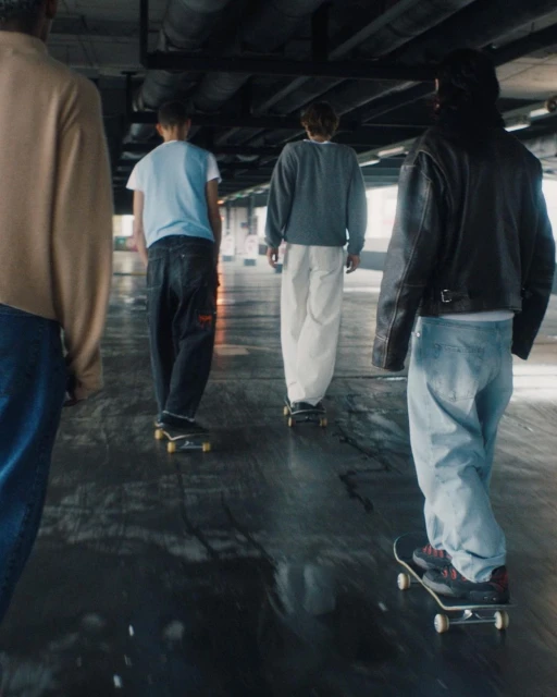 Group of teens skateboard on a wet, urban surface in an underpass, showing casual clothing, skateboards, and movement with a wide perspective.