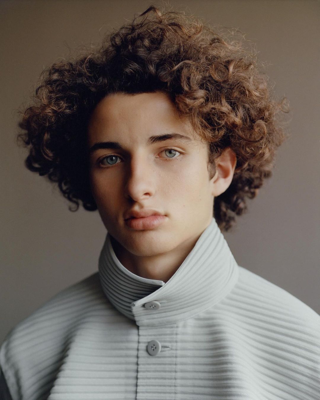 Close-up portrait of a young adult male with curly brown hair wearing a light blue ribbed jacket, photographed indoors against a neutral background.