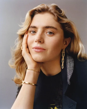 Close-up portrait of a woman resting her chin on her hand, wearing gold chain earrings, bracelets, and a ring.