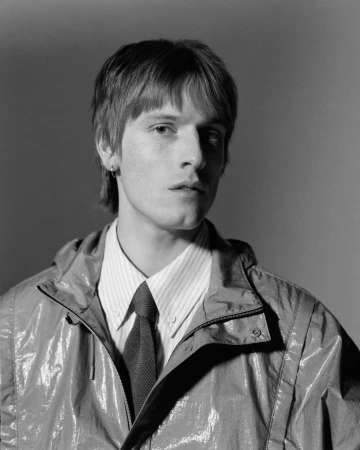 Portrait of a man wearing a hooded jacket, striped button-down shirt, and tie against a plain grey studio background.