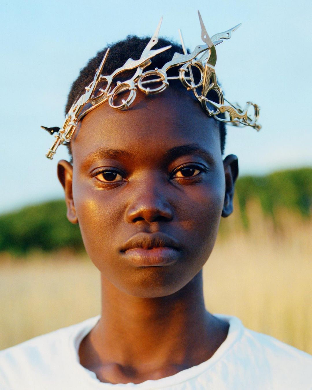 Close-up portrait of a young Black female wearing a crown made of scissors, posing outdoors with a neutral expression and soft, natural light.