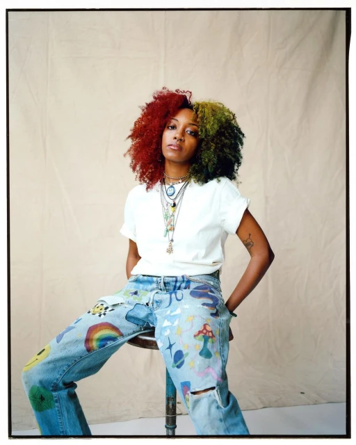 An adult female sits on a stool wearing custom-painted jeans, a white t-shirt, and multiple necklaces against a neutral background.