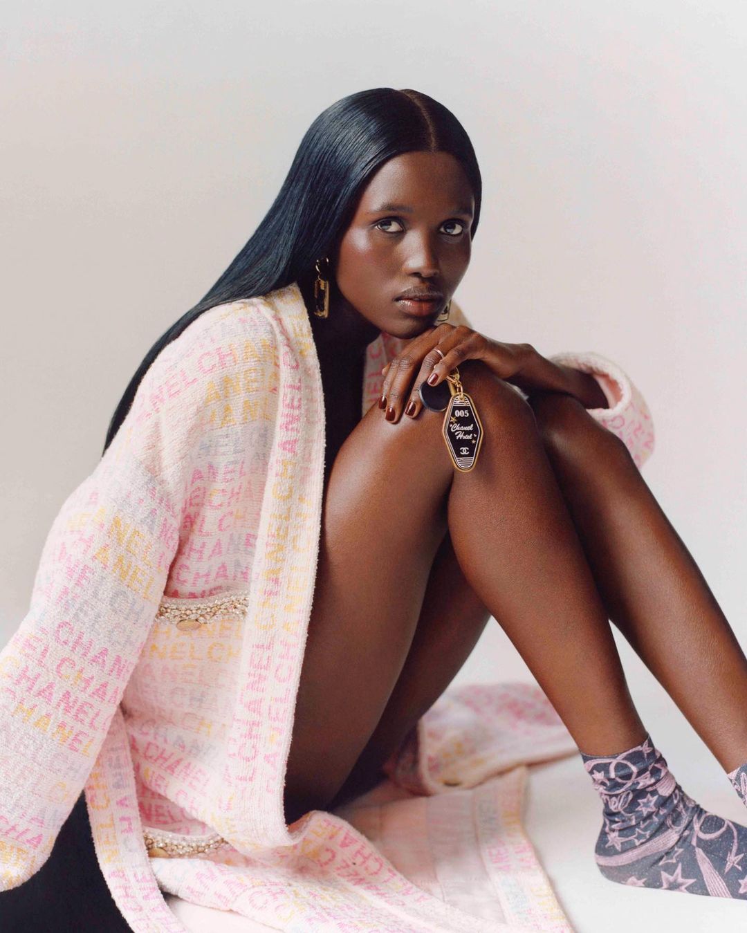 An elegant Black woman models a pastel robe with Chanel branding while holding a Chanel logo keychain in a studio setting.