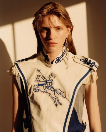 Portrait of a female model wearing an embroidered cream and blue military-style vest against a plain wall.