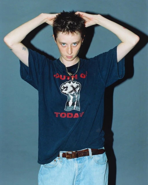 A person with short brown hair and wearing a black t-shirt with a fist graphic poses against a blue-grey backdrop, hands raised.