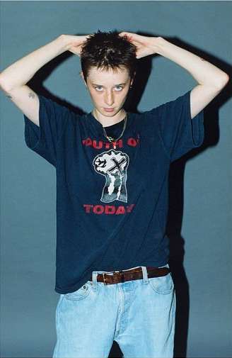 A person in a dark T-shirt with graphic print and light blue jeans stands in front of a grey backdrop, posing with hands above their head.