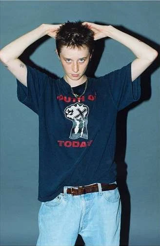 A person in a dark T-shirt with graphic print and light blue jeans stands in front of a grey backdrop, posing with hands above their head.
